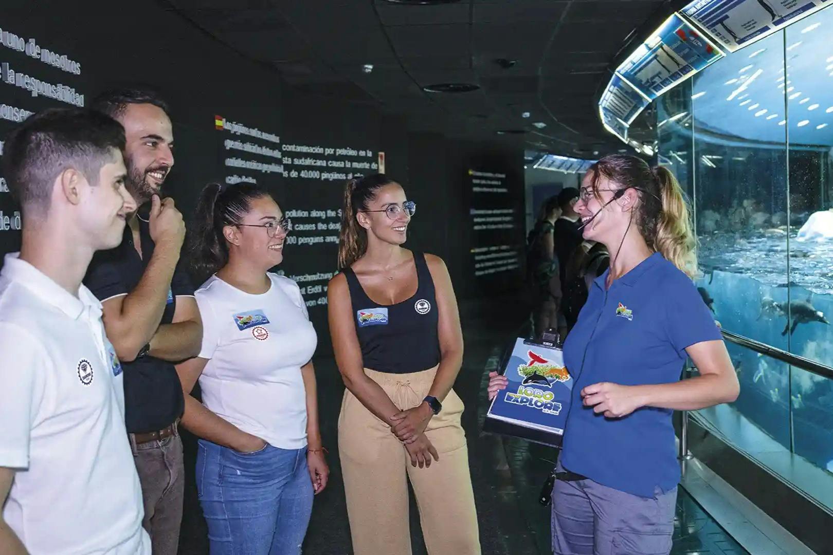 Four people stand indoors, smiling and talking near a display with text. They wear casual clothing with badges.