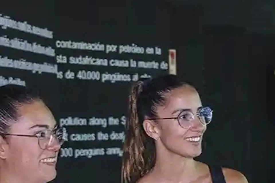 Four people stand indoors, smiling and talking near a display with text. They wear casual clothing with badges.
