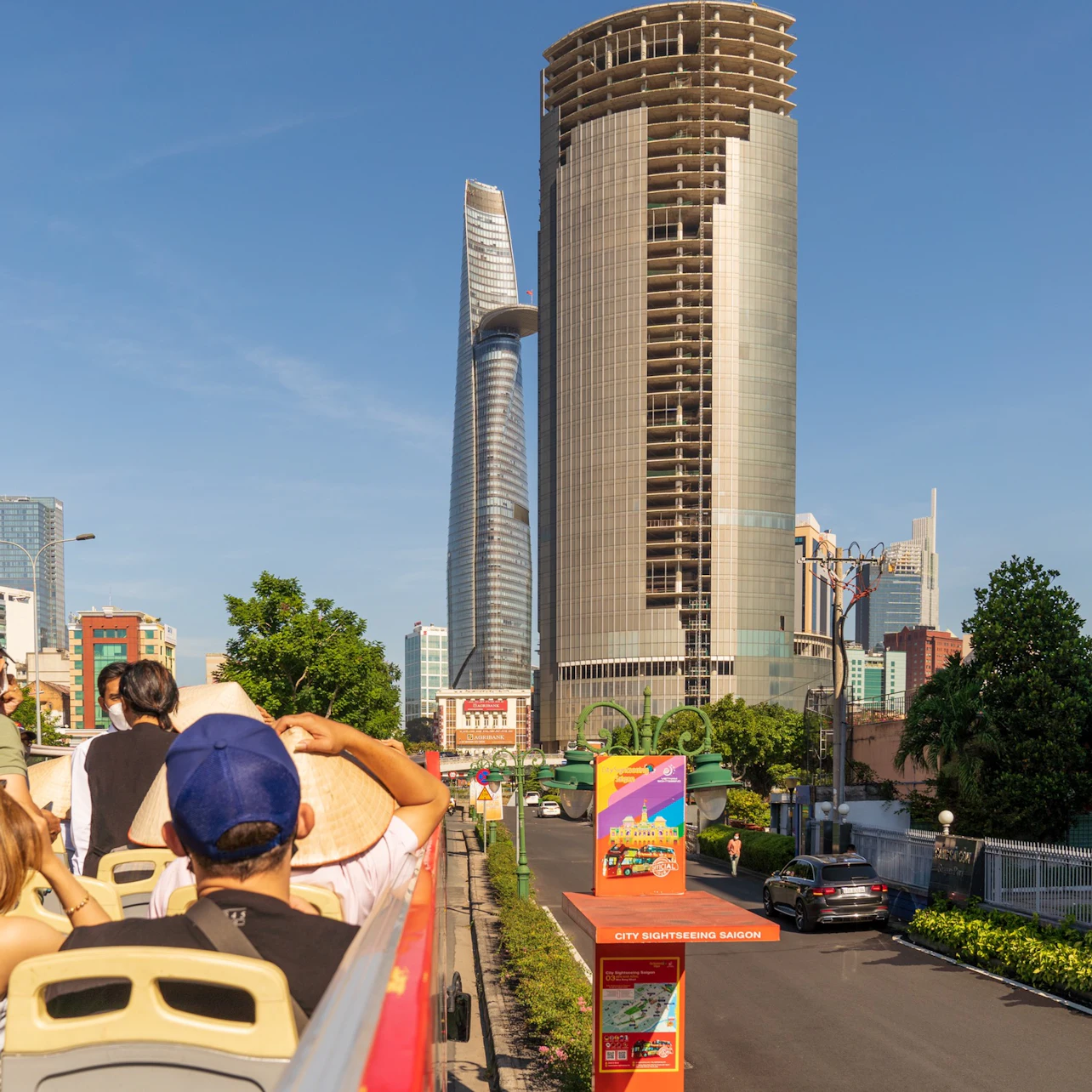 A group of people on a double-decker bus touring a city with tall buildings and greenery under a clear blue sky.