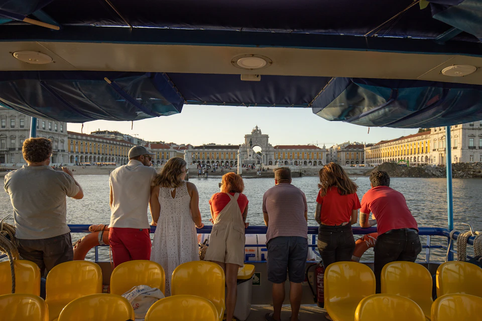 People stand on a boat, looking at a large, yellow waterfront building with a central arch in the distance.