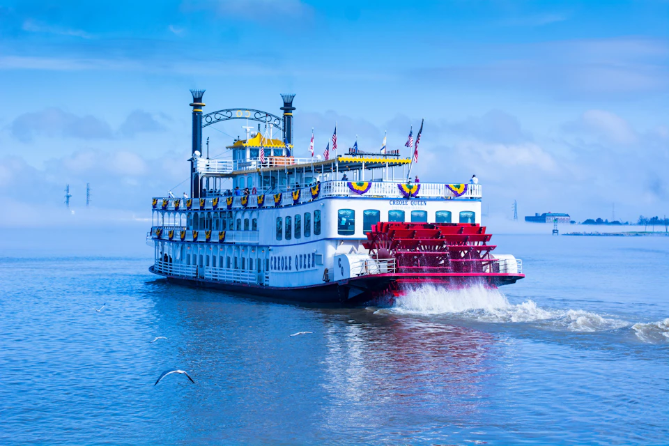 A decorated paddle steamboat, "Creole Queen," on blue water with flags and yellow-blue banners, under a clear sky.