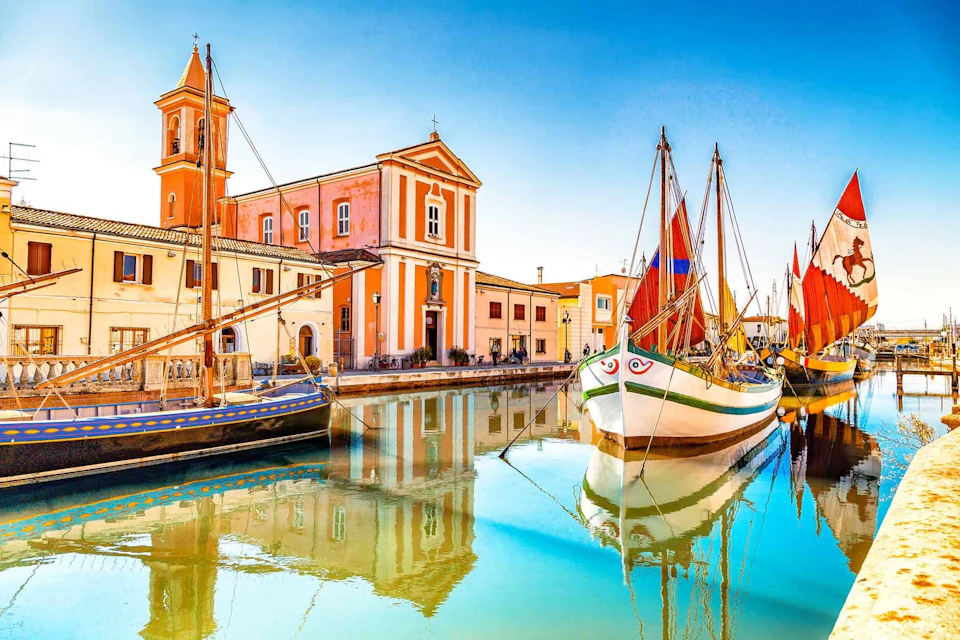 Colorful sailboats docked beside vibrant buildings with a church tower, reflected in calm canal water under a clear blue sky.