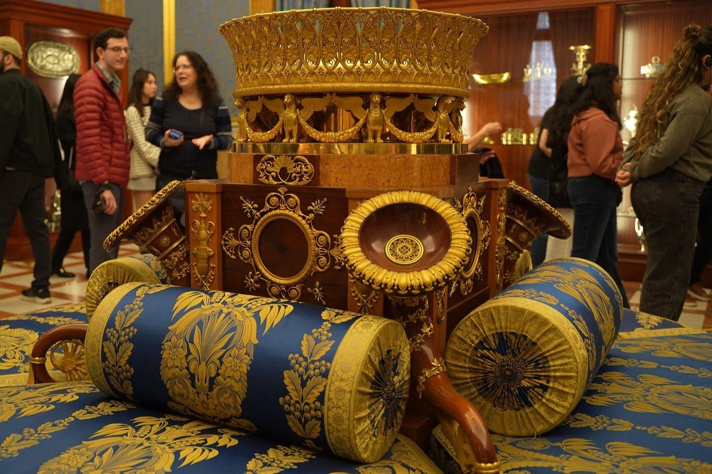 A detailed ornate piece of furniture with gold and blue decorations, surrounded by people in a museum-like setting.