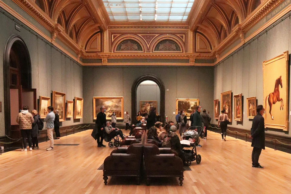 People observe paintings in a spacious art gallery with a high, ornate ceiling and cushioned benches in the center.