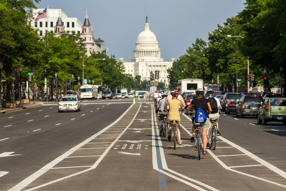 People riding bicycles in a bike lane towards the U.S. Capitol building on a sunny day. Traffic and trees line both sides of the street.