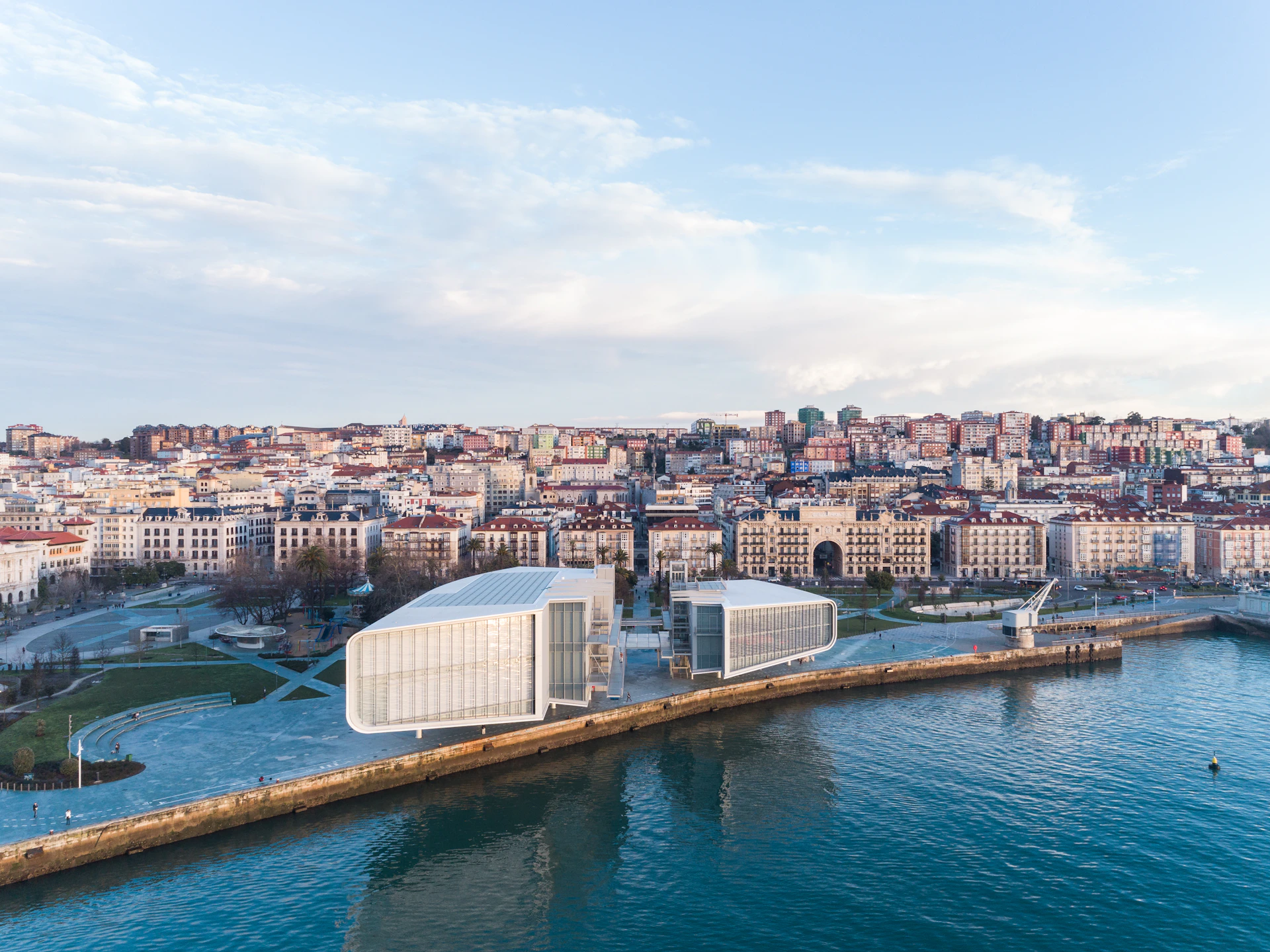 Modern waterfront buildings near a cityscape with densely packed buildings, a park area, and a clear blue sky.