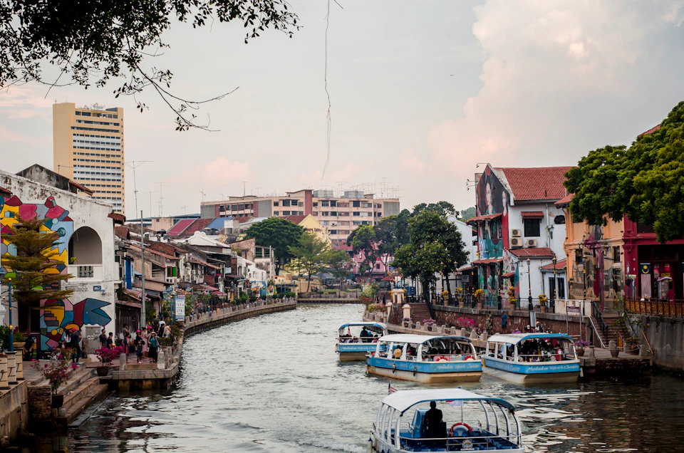 Several tour boats navigate a river through an urban area with trees and buildings, while people walk along the riverside walkway.