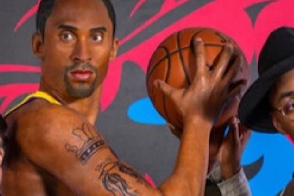 Two men pose with a wax figure of a basketball player in a yellow jersey, set against a colorful background.
