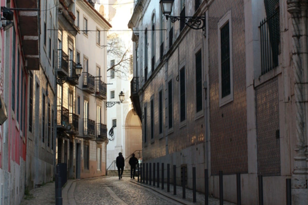 两个人走在一条狭窄的鹅卵石街道上，两侧是带阳台和路灯的高楼大厦。