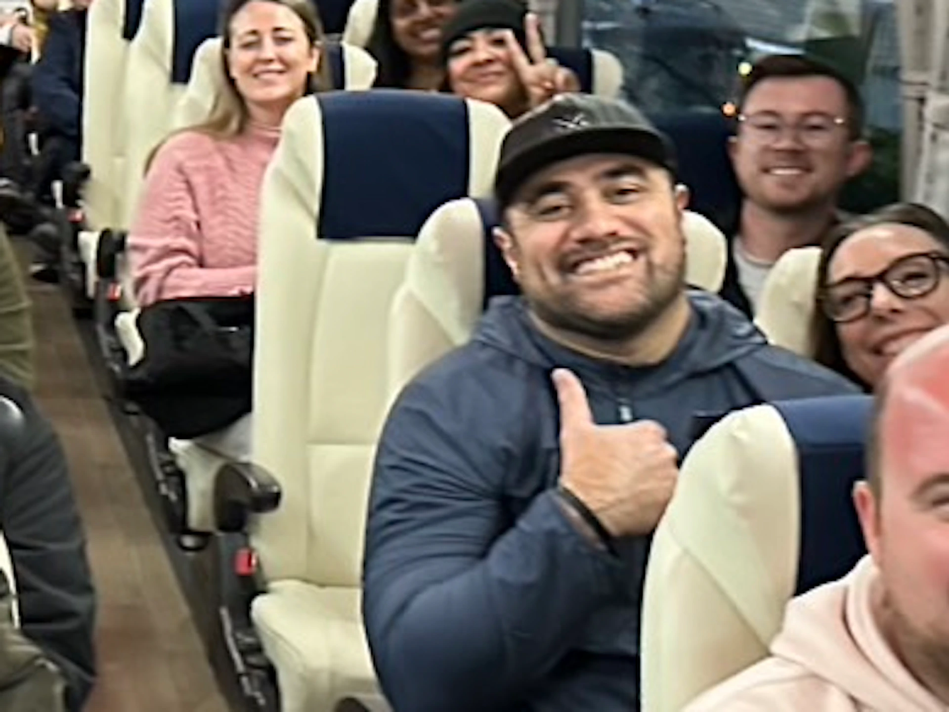 A group of people seated inside a well-lit bus, smiling towards the camera, with red lighting accents on the ceiling.