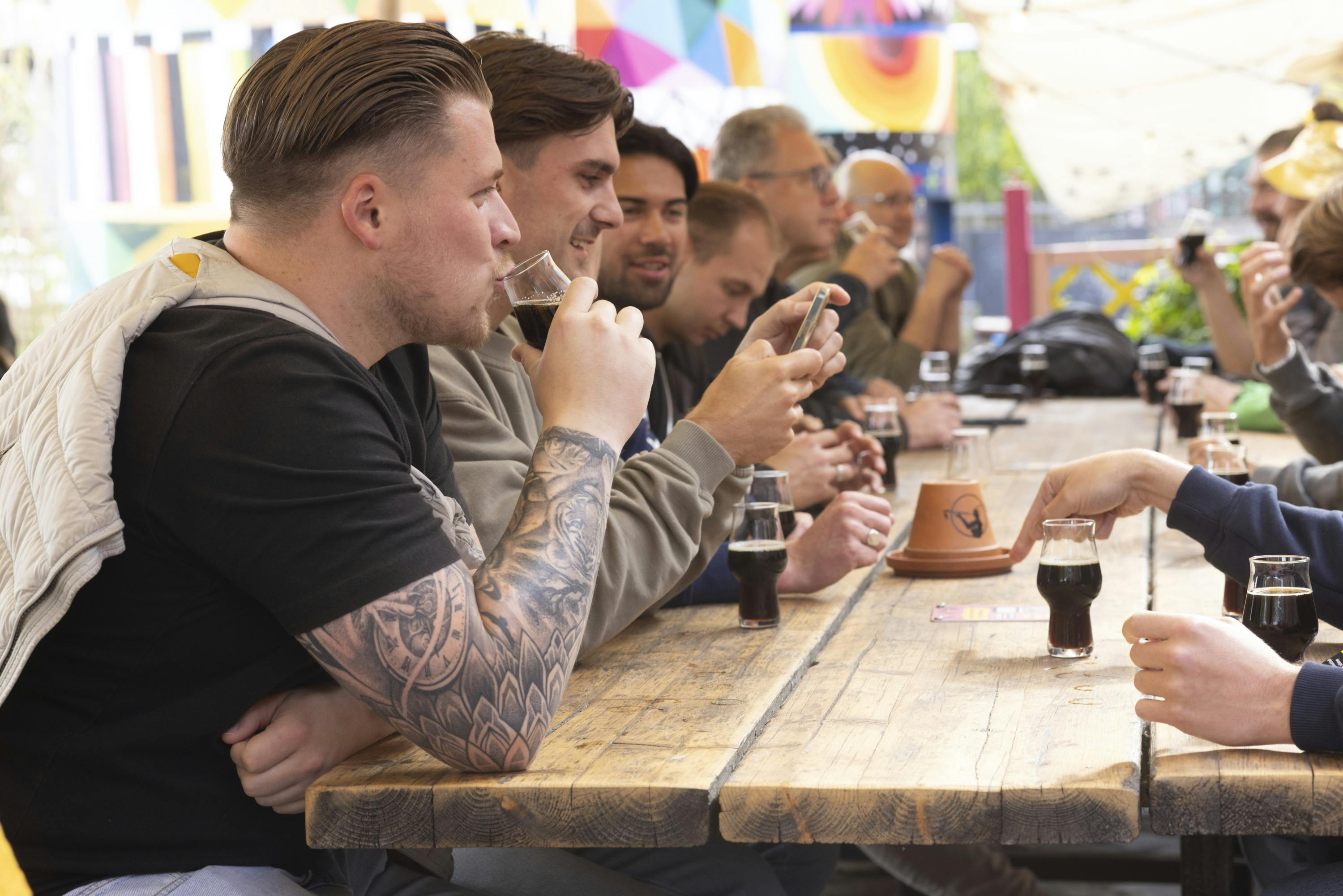 En gruppe mennesker, der sidder ved et langt træbord og drikker og hygger sig. Manden i fokus har tatoveringer, nogle ser på telefoner. Dekoreret baggrund.