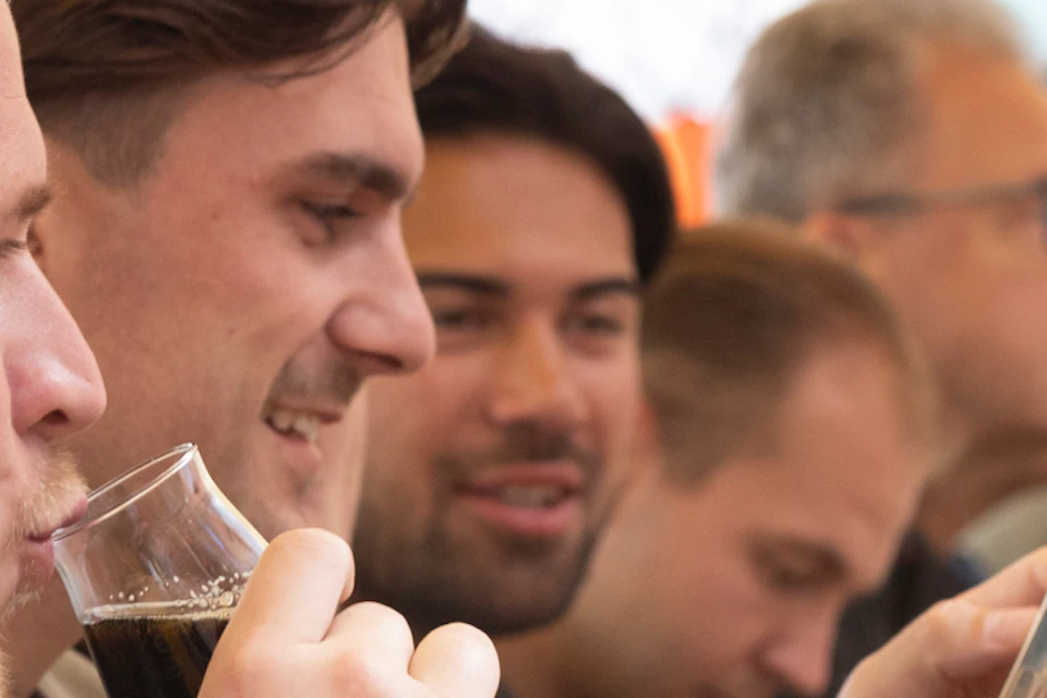 Group of people sitting at a long wooden table, drinking and socializing. Man in focus has tattoos, some look at phones. Decorated background.