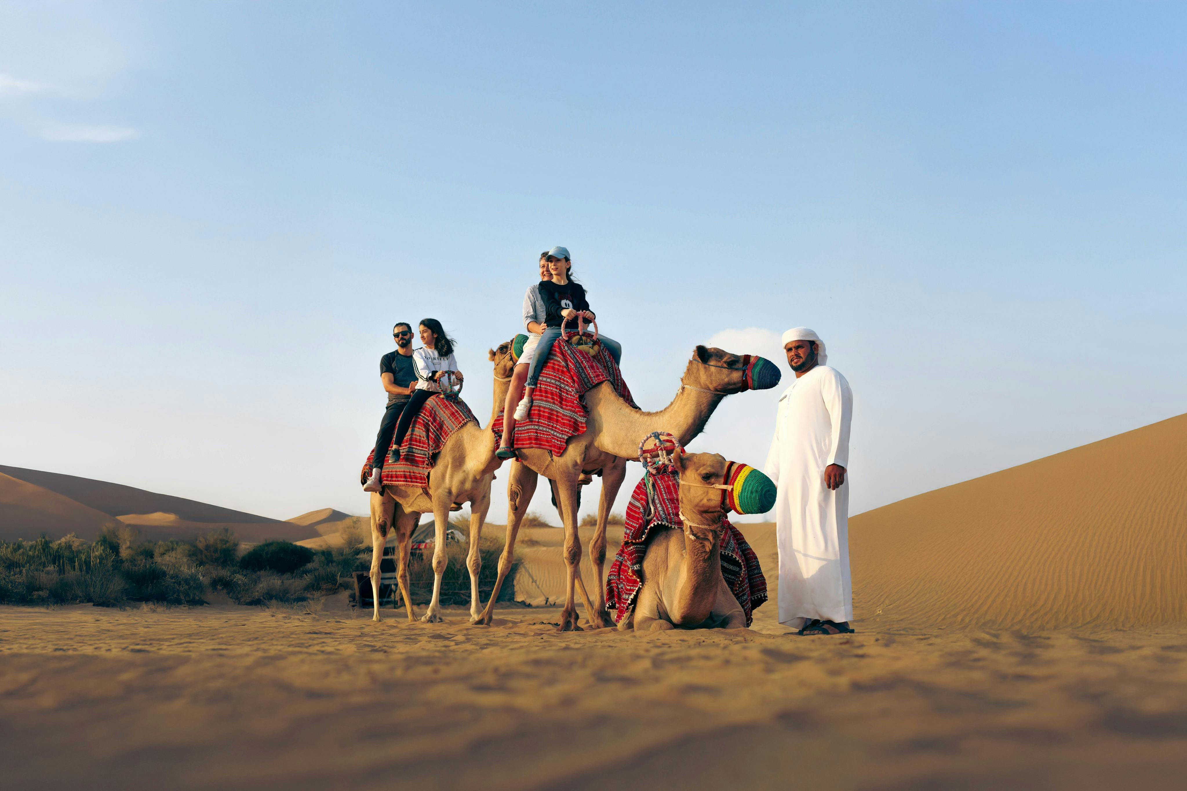 Menschen reiten auf mit bunten Decken geschmückten Kamelen in der Wüste, geführt von einem Guide in weißer Tracht. Sanddünen und blauer Himmel im Hintergrund.