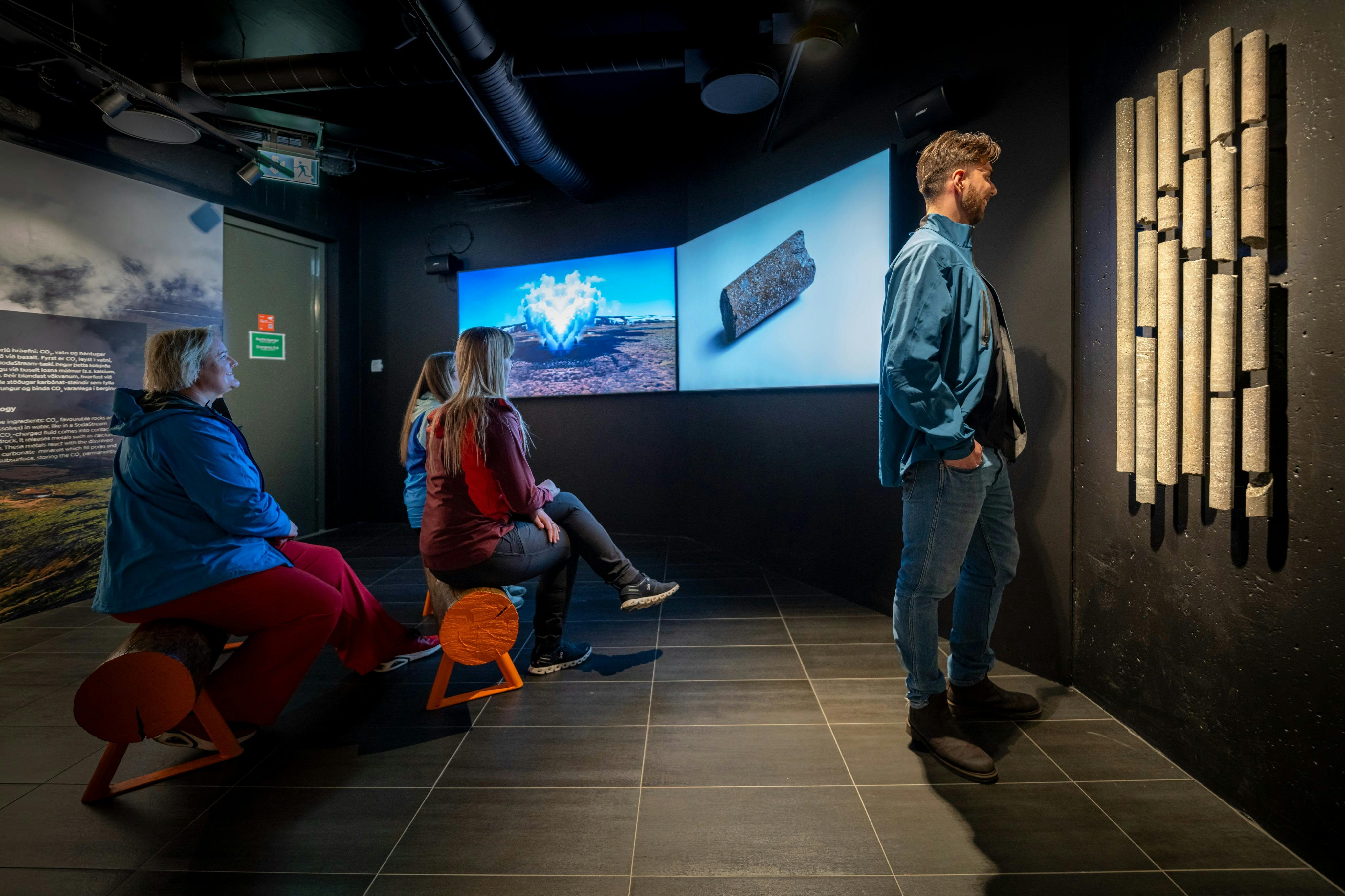 Menschen in einem schwach beleuchteten Raum, die große Bildschirme mit Bildern von Landschaften und Objekten betrachten, mit moderner Einrichtung und Sitzgelegenheiten.