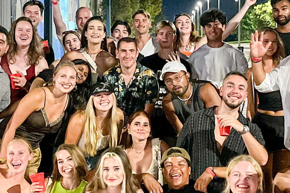A group of people posing together outdoors at night, some holding drinks, with a lit street and trees in the background.
