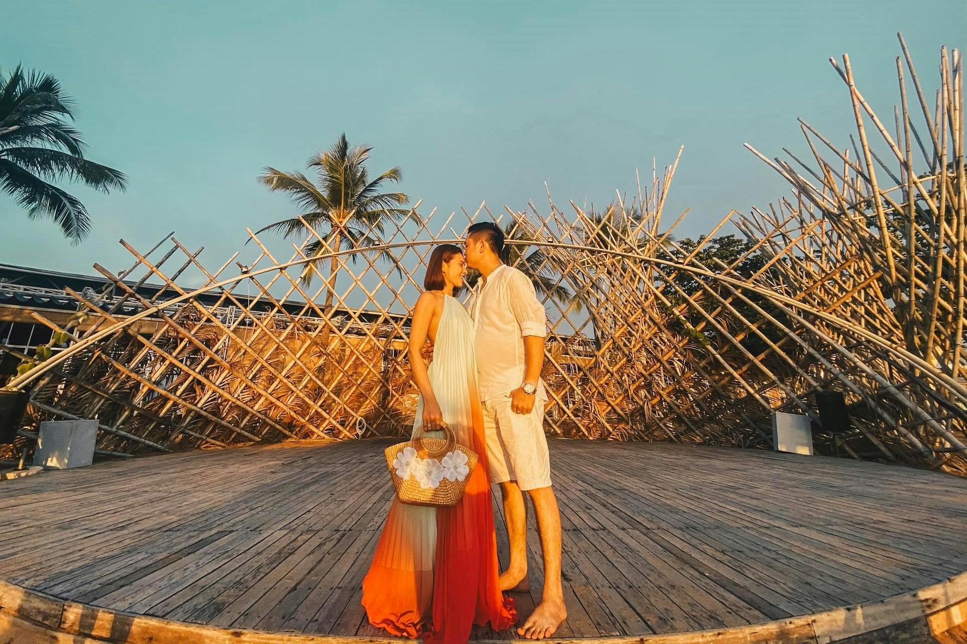 A couple stands on a wooden platform with a bamboo structure and a palm tree in the background, under a clear sky.