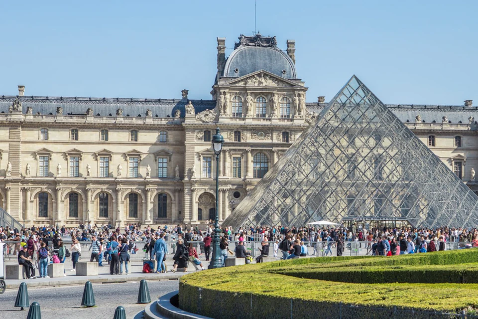 A crowd of people outside the Louvre Museum in Paris, featuring the glass pyramid and historic building on a clear day.