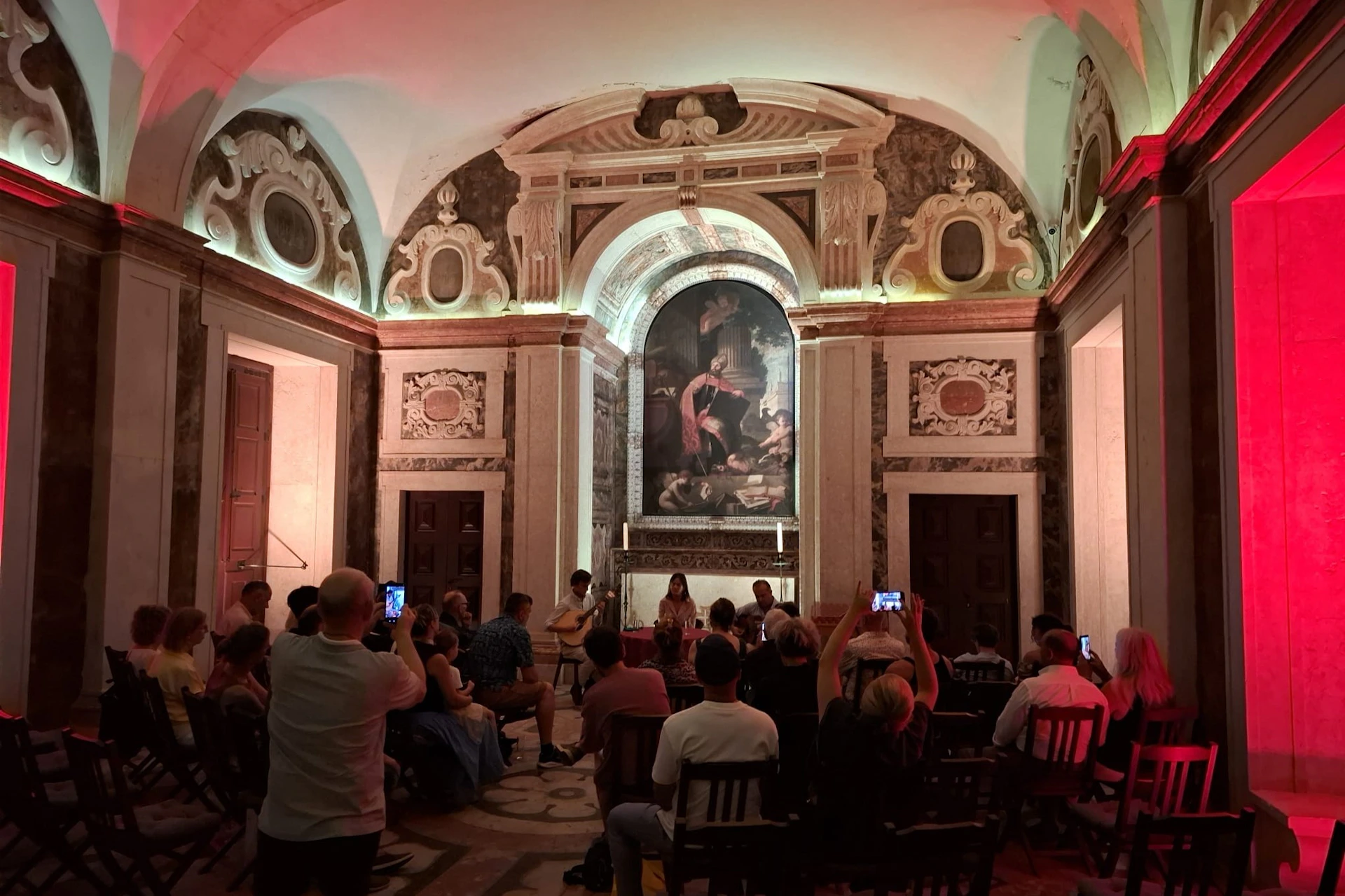 A group of people seated in a decorated hall with an archway and a large painting, during a performance or event.