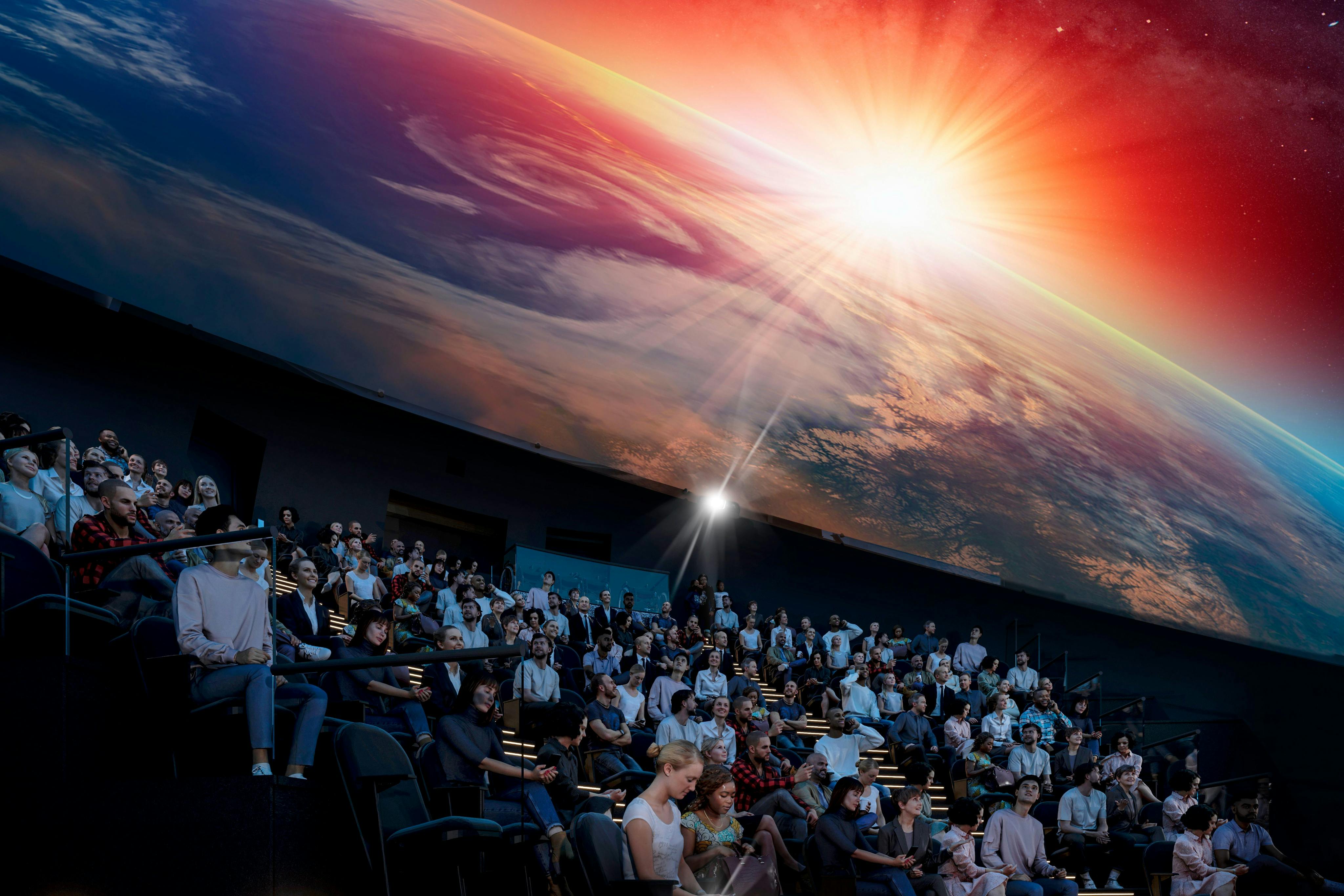 Velké publikum sedí v planetáriu a pozoruje živou projekci západu slunce nad Zemí.