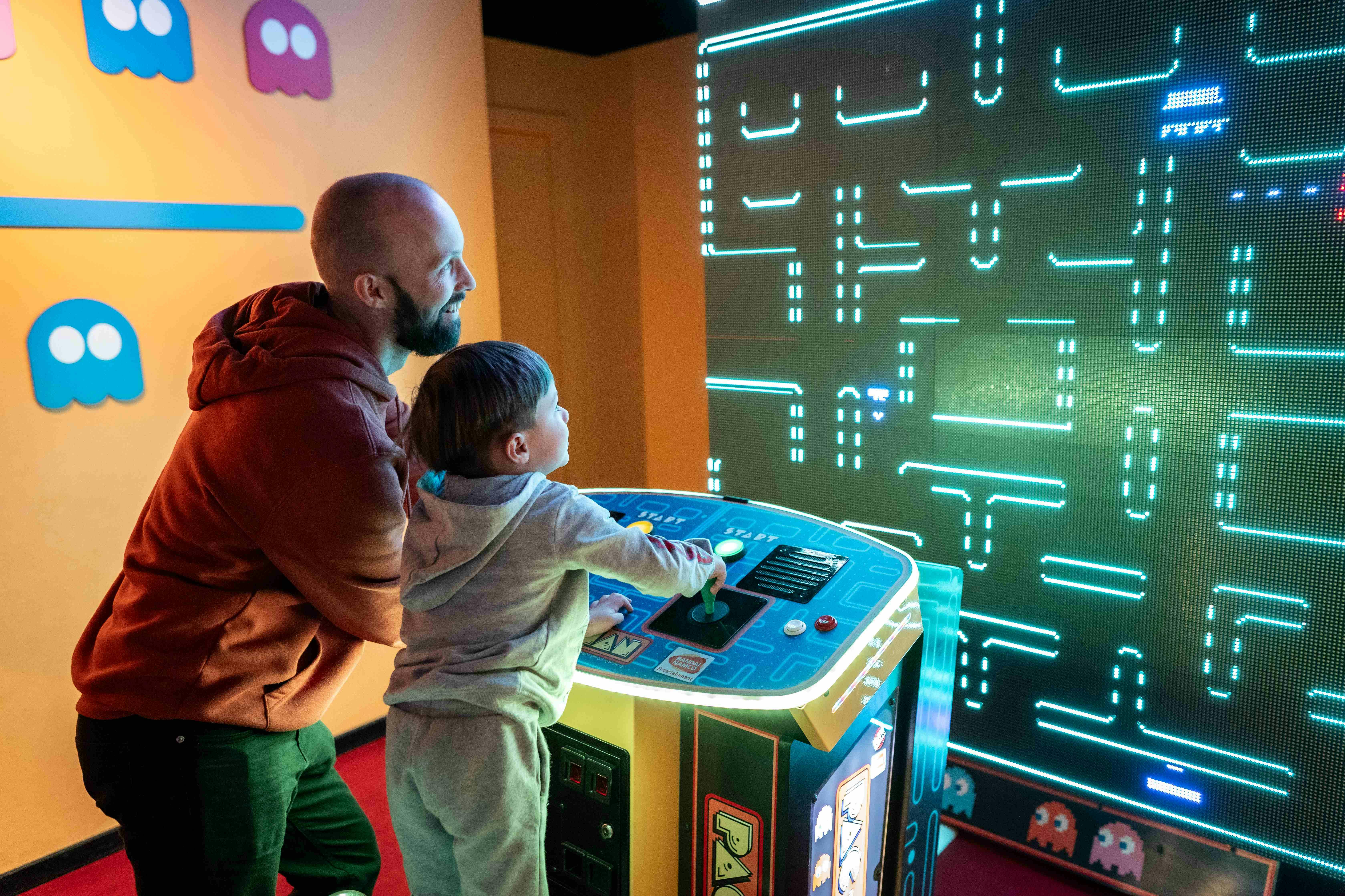 A man and a child play on a large retro arcade machine, with colorful ghost characters on the wall behind them.