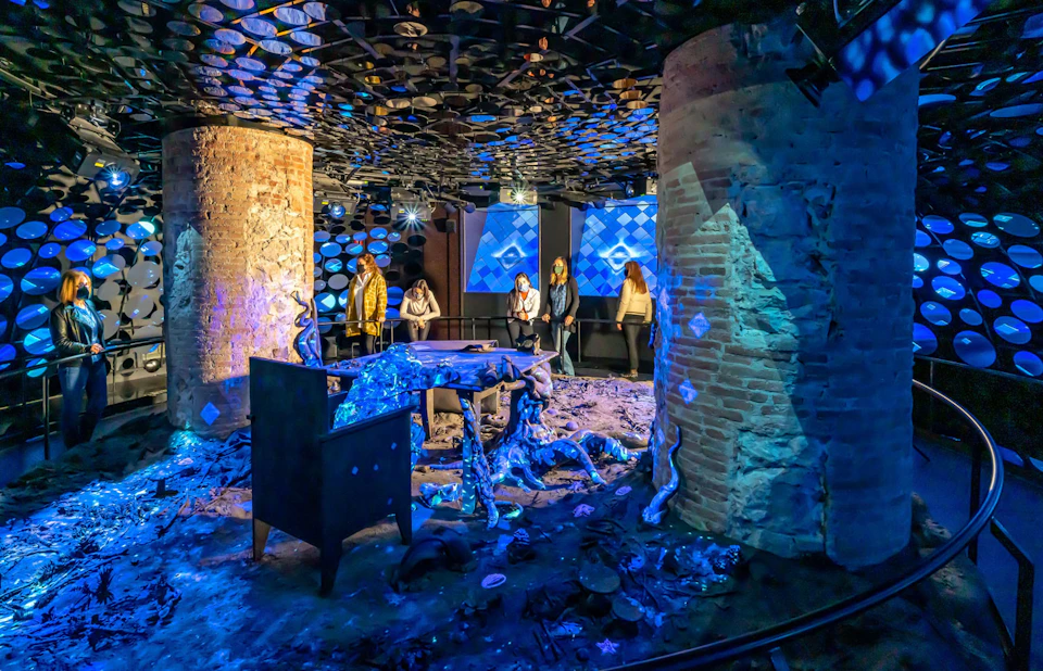 People observe an art installation with blue lighting, featuring brick columns, organic shapes, and reflective circular objects.