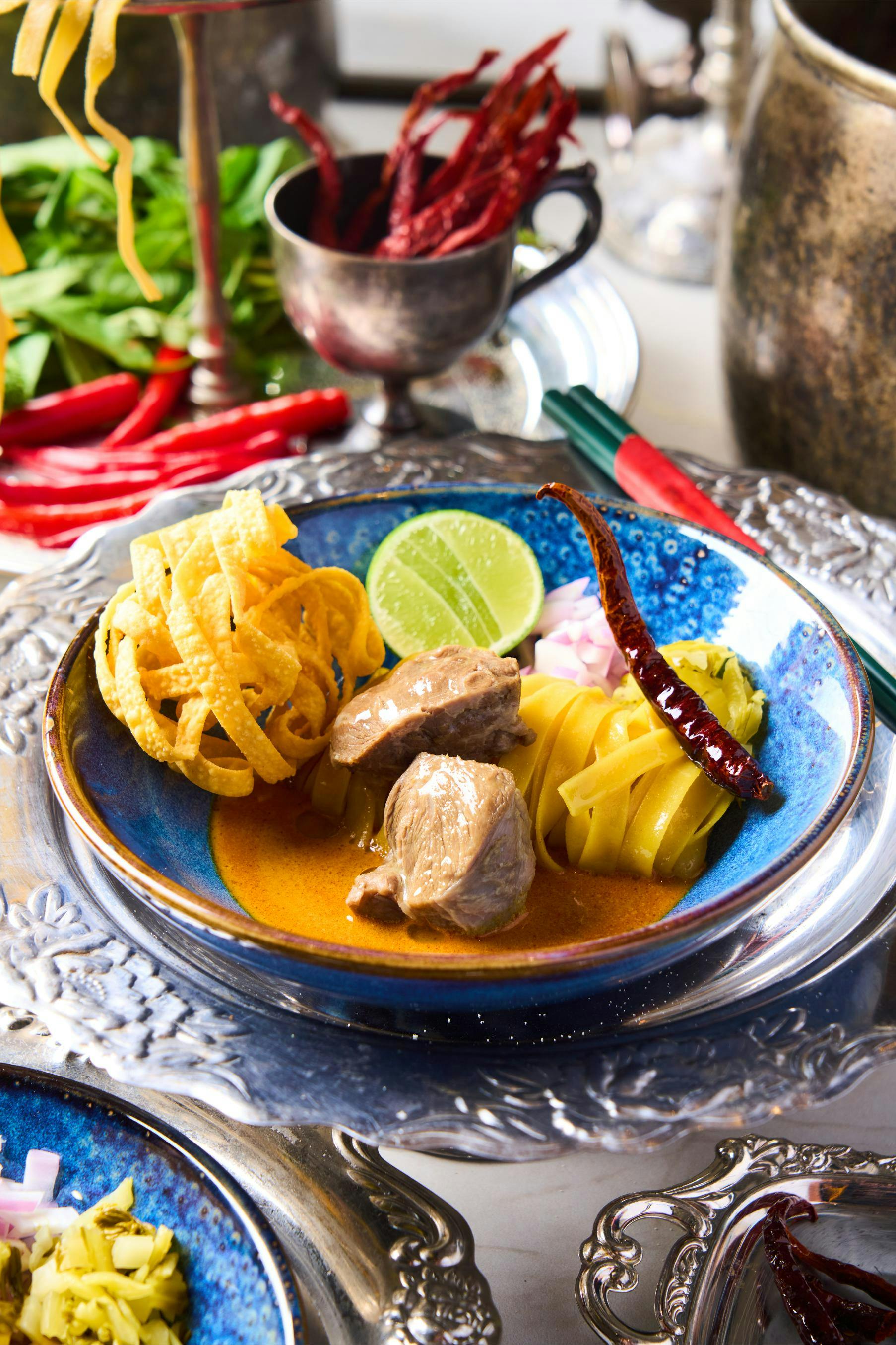 A bowl of curry with meat, noodles, garnishes, and lime, served on a decorative metallic plate with utensils nearby.