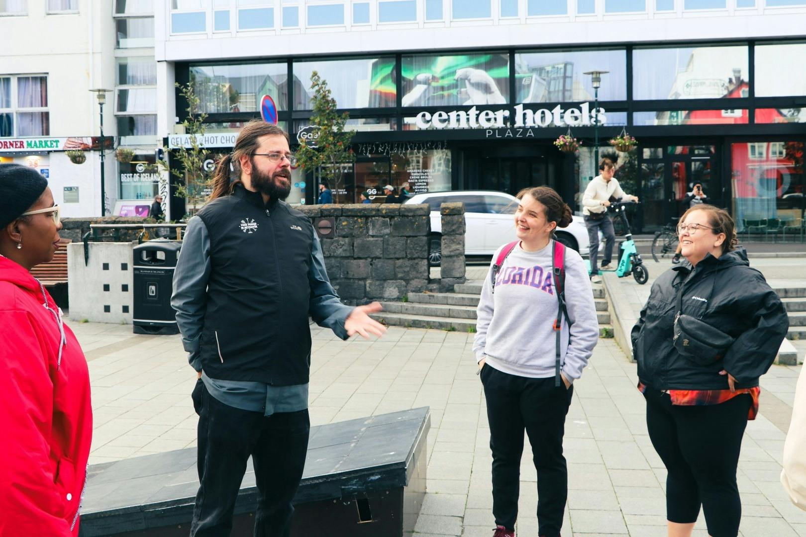 세 명의 일행이 도심의 야외에 서서 대화를 나누고 있습니다. 돌담과 건물이 배경입니다.