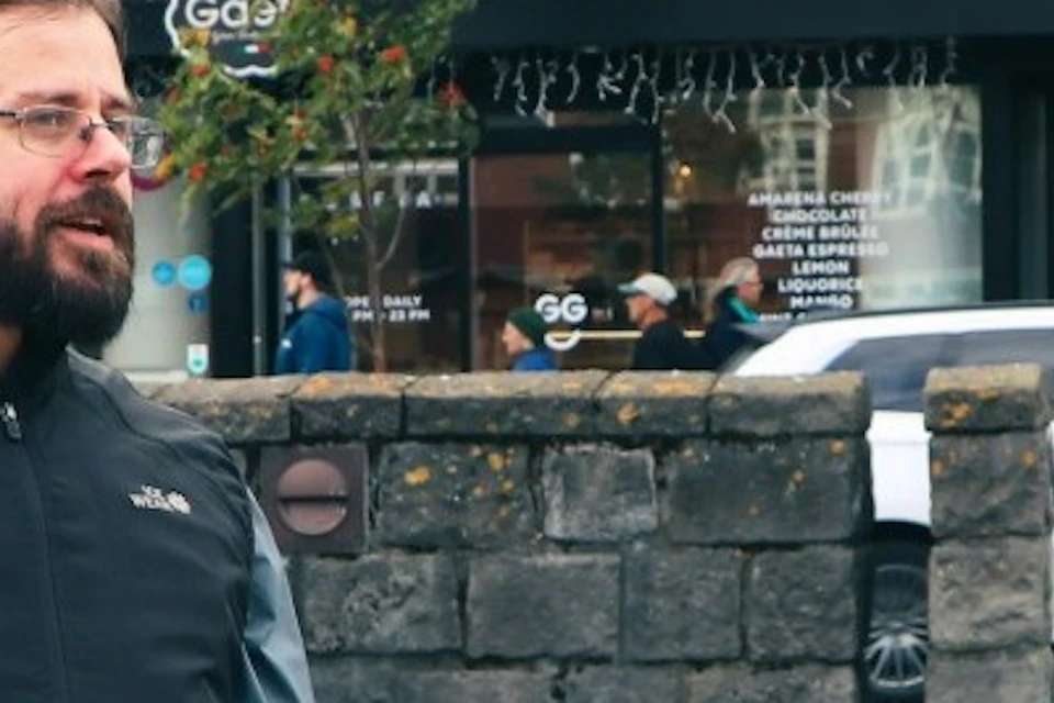 A group of three people stand in an urban outdoor setting, engaged in conversation. A stone wall and buildings are in the background.