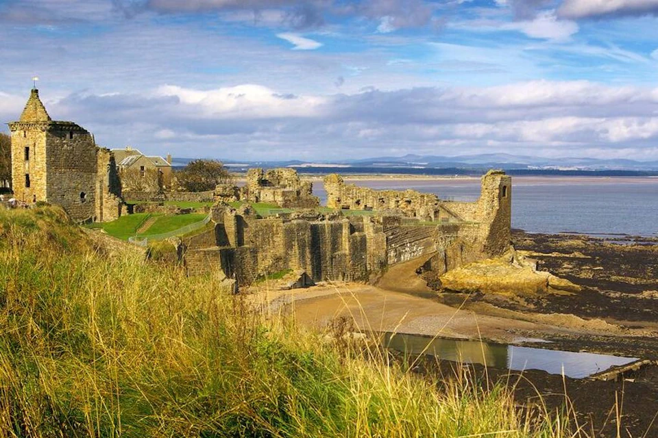 Ruins of a historic stone castle overlooking a coastal area with grass, a sandy shore, and a distant horizon under a cloudy sky.
