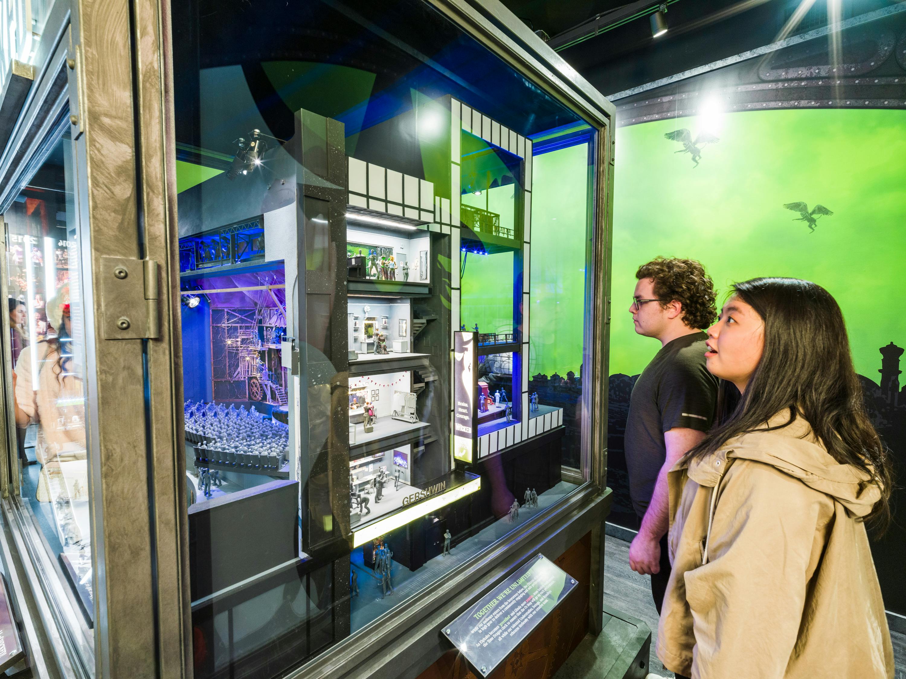 Δύο άτομα παρατηρούν ένα λεπτομερές, πολυεπίπεδο διόραμα σε ένα μουσείο με πράσινο φόντο που εμφανίζει ιπτάμενα πλάσματα.