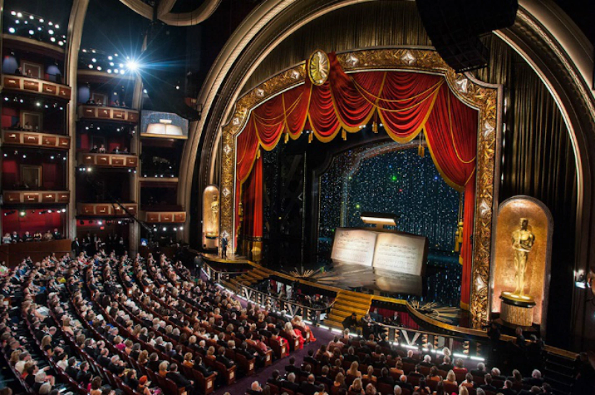 Elegant theater filled with seated audience; stage with open book prop, red curtains, and ornate gold arch.