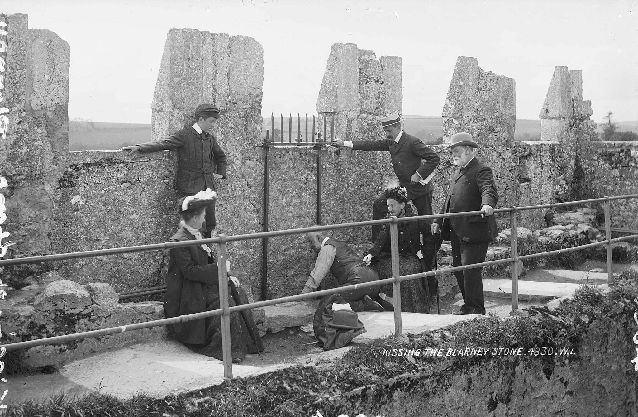 A group of people gathered around a man kneeling and leaning backward to kiss the Blarney Stone at a stone wall site.