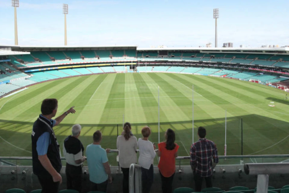 A group of people standing in a stadium, viewing a large green field with goal posts under a clear sky.
