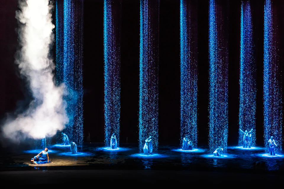 Illuminated performers in blue light under cascading water streams on a dark stage with mist to the side.