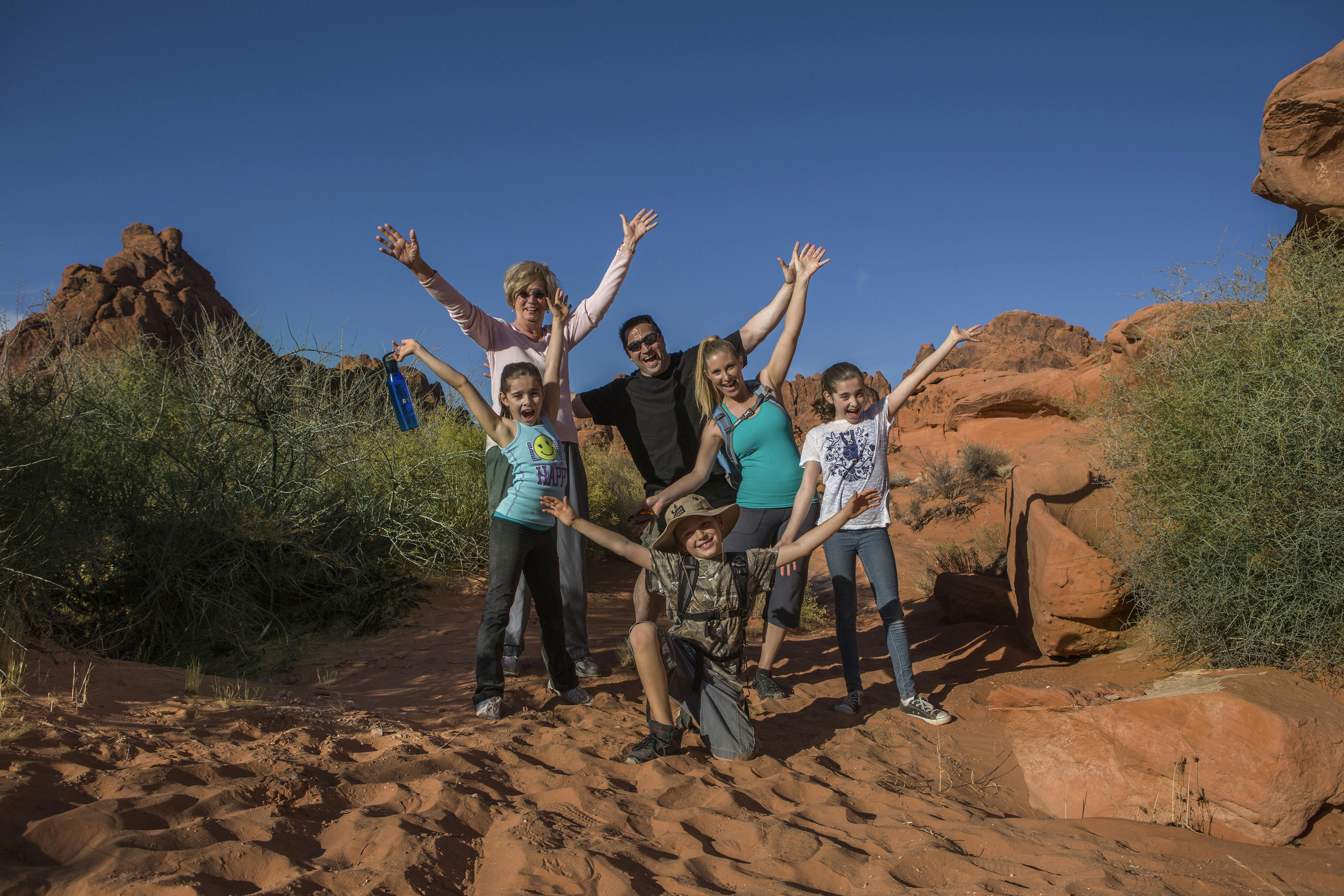 Family of six joyfully posing with arms raised in a desert setting, surrounded by red rocks and sparse vegetation.