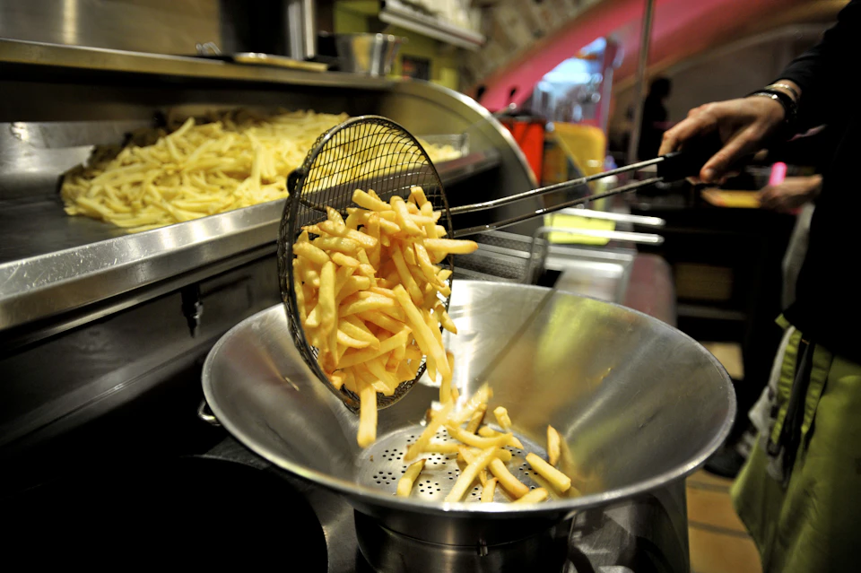 Freshly cooked French fries being lifted from a fryer with a strainer into a metal bowl, with more fries in the background.