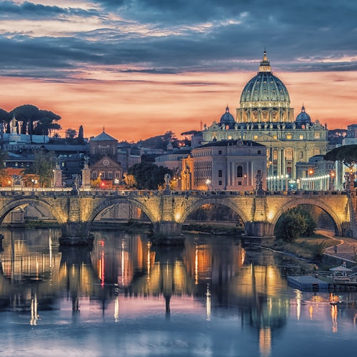 View of a lit-up bridge and St. Peter's Basilica at sunset with reflections in the river below.