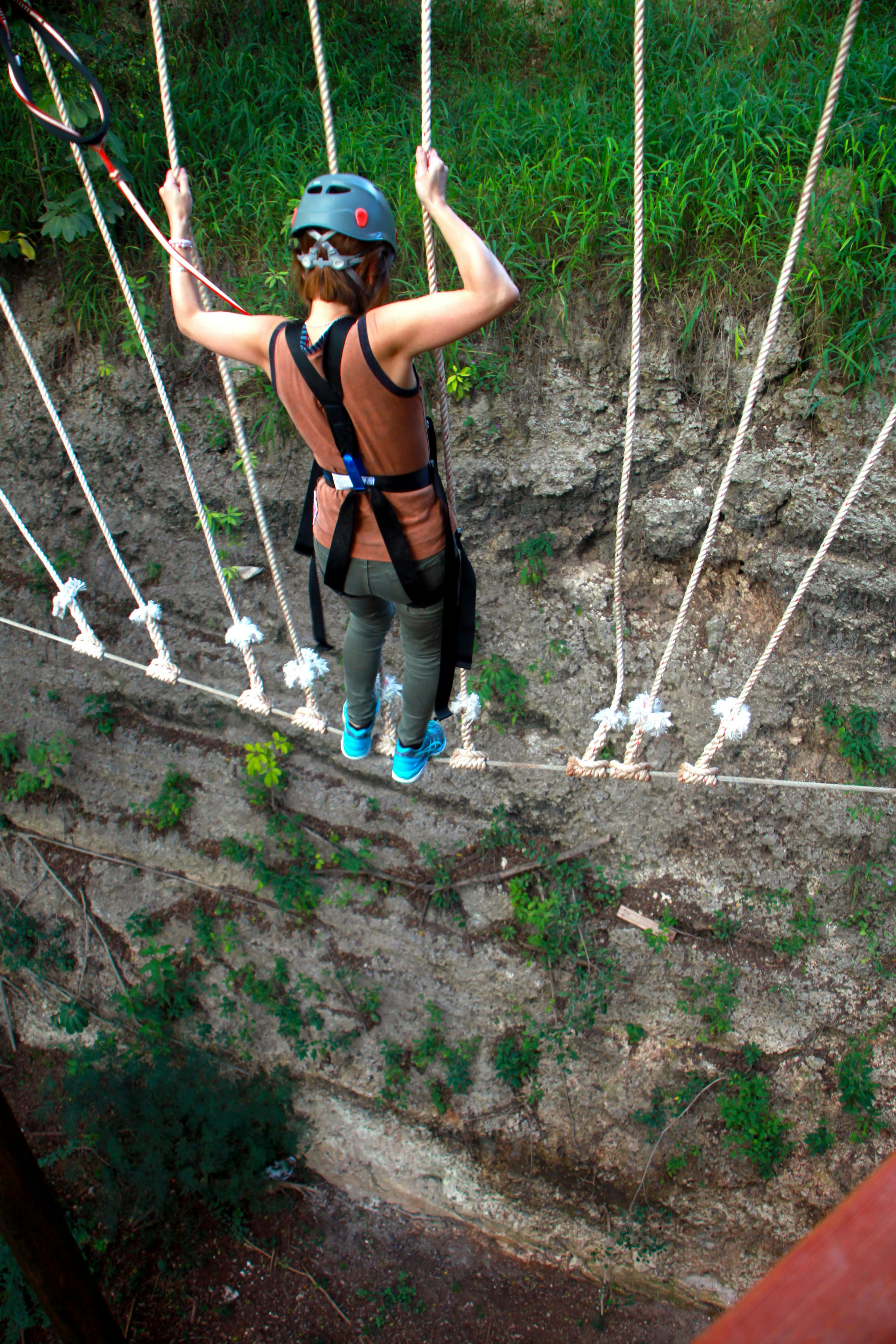 아래 바위와 초목이 있는 가파른 협곡 위로 밧줄다리를 건너는 하네스를 입은 사람.