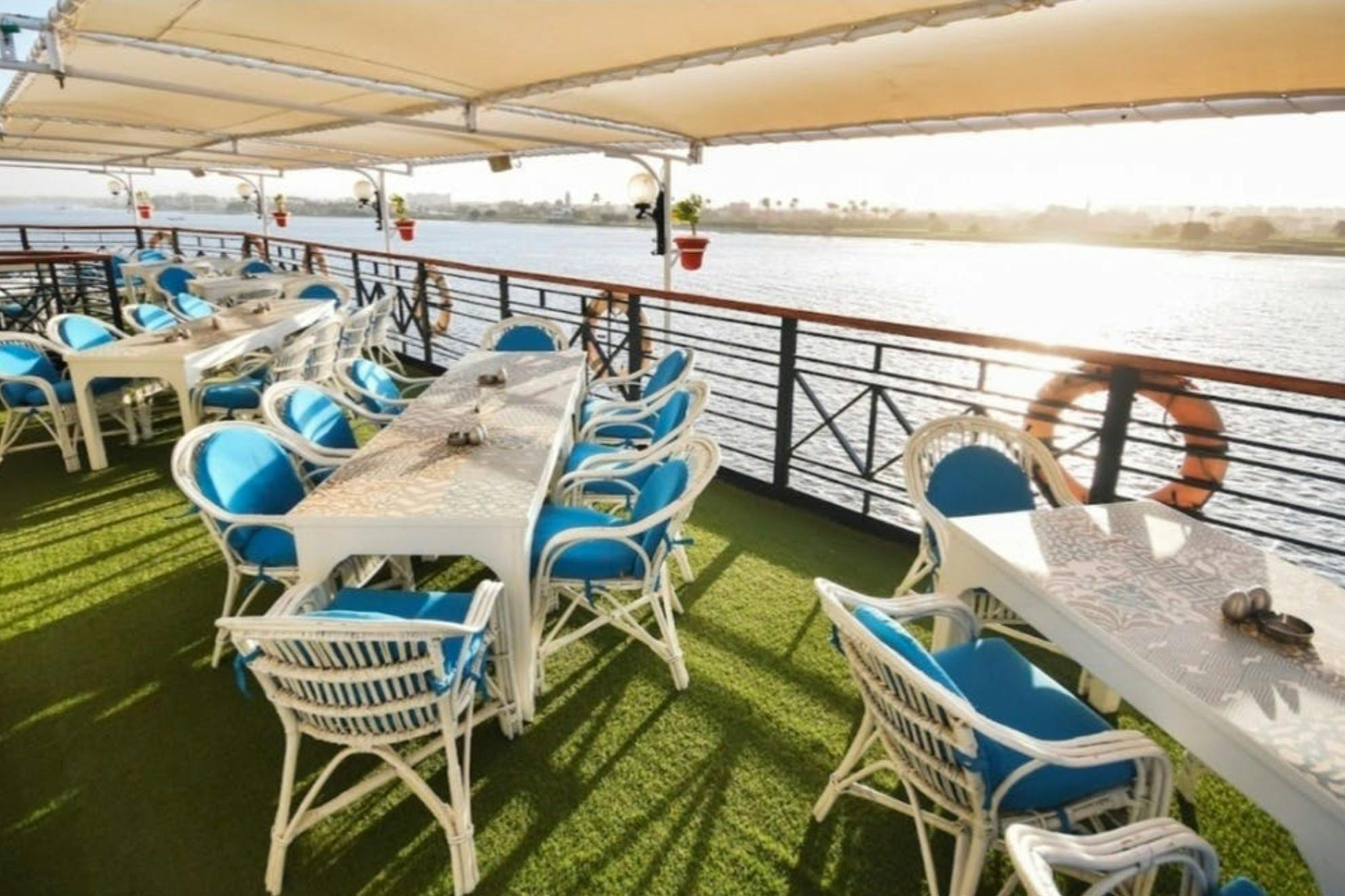 Ein Sitzbereich im Freien auf einem Boot mit weißen Tischen, blauen gepolsterten Stühlen und Blick auf einen Fluss unter einem Baldachin.