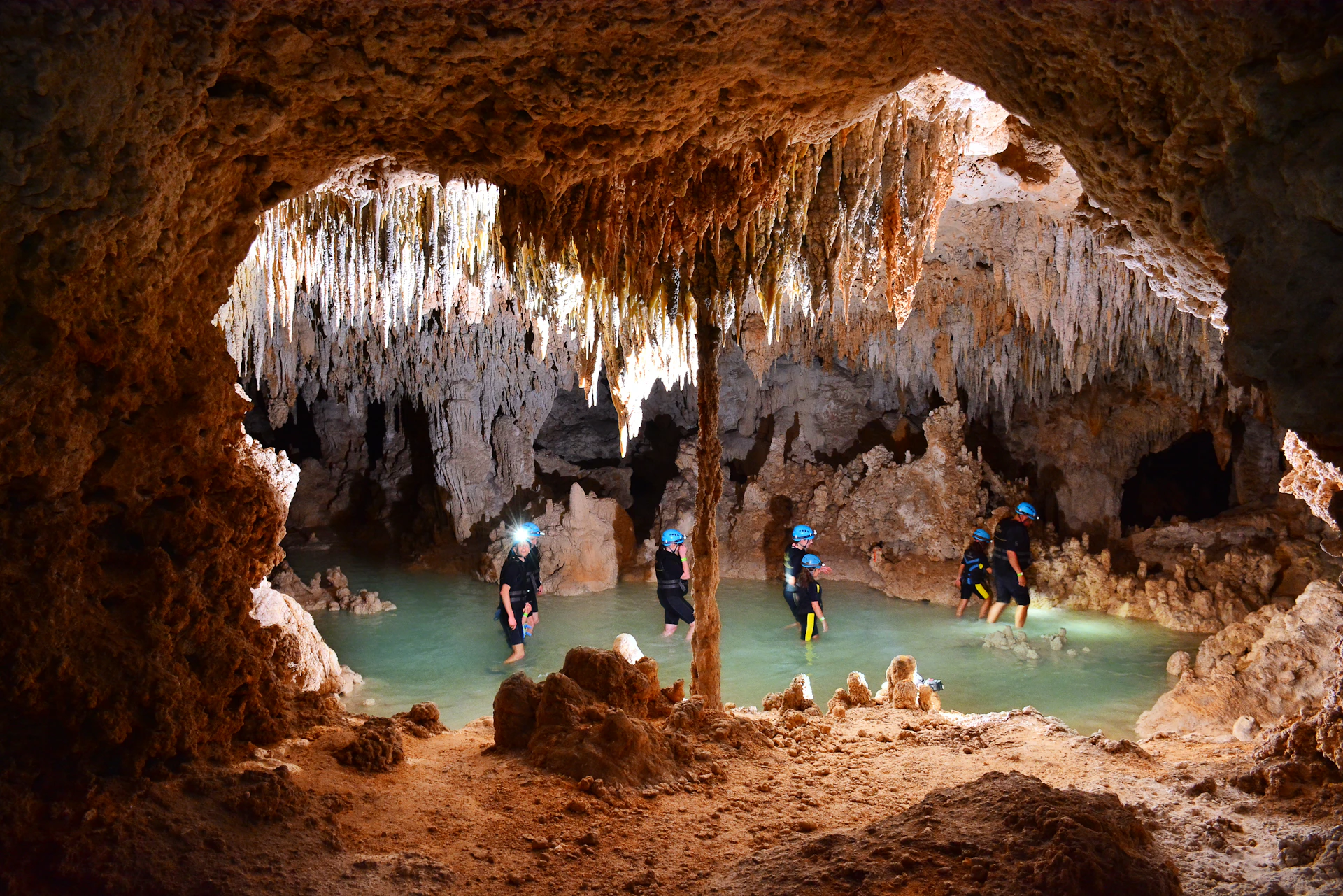 People wearing helmets and headlamps wade through waist-deep water inside a cave with numerous stalactites and stalagmites.
