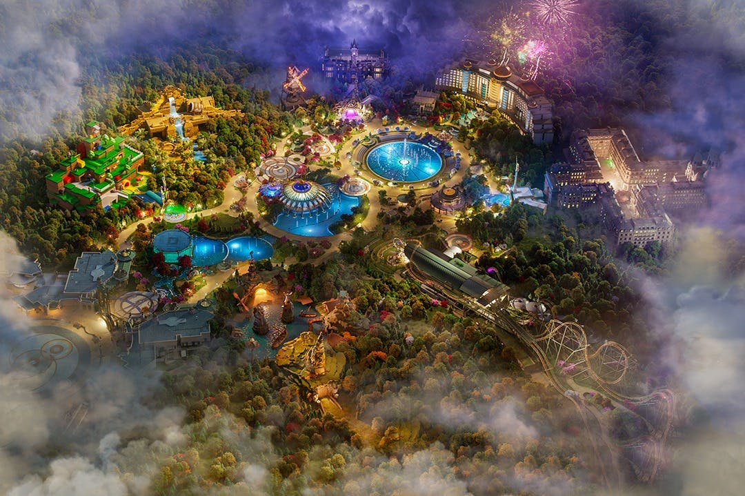 Vista aerea di un parco divertimenti con giostre, alberi, giochi d'acqua, edifici illuminati e fuochi d'artificio nel cielo.