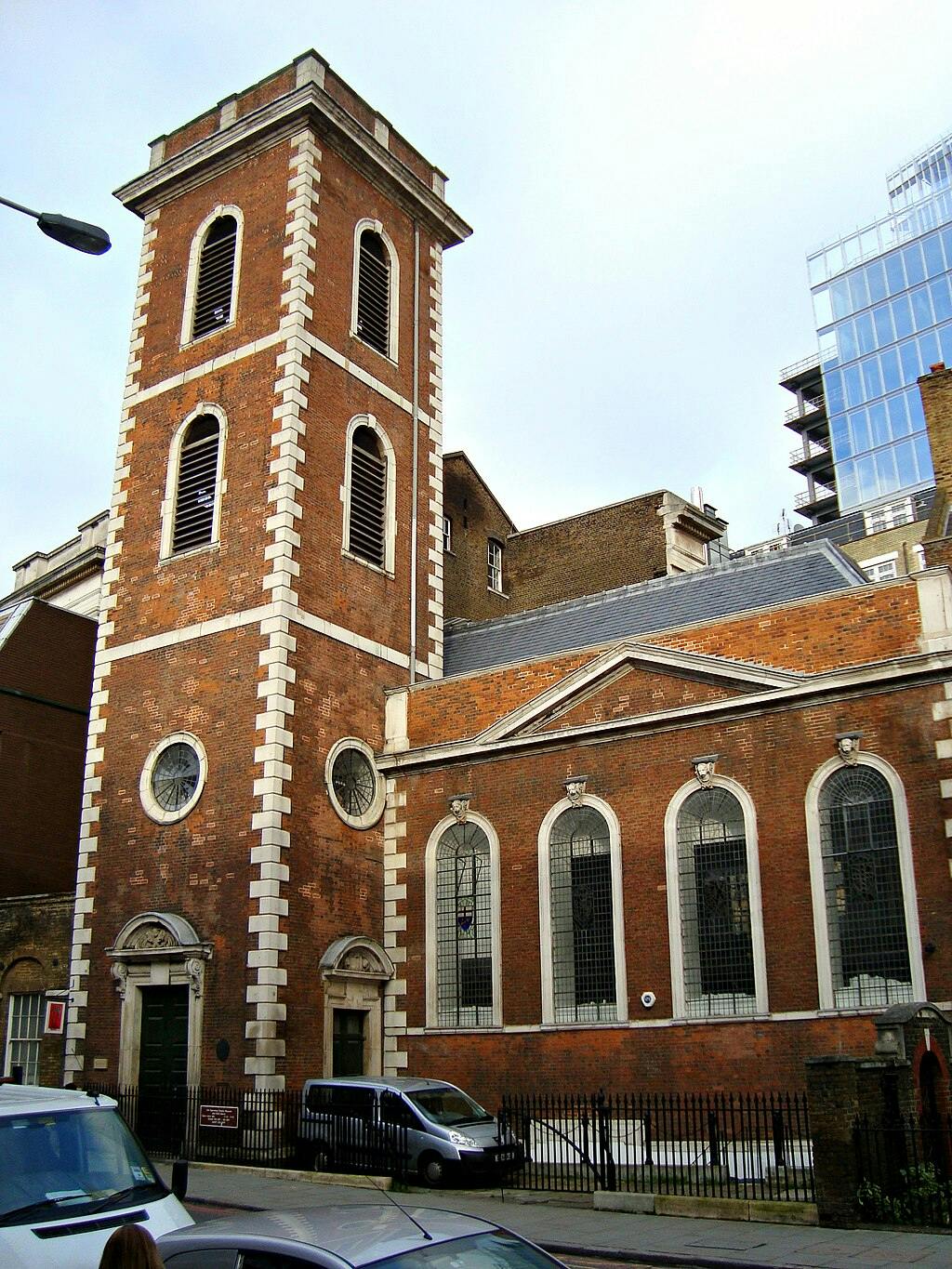 Murstensbygning med højt tårn, buede vinduer og en indhegnet indgang på en bygade med parkerede biler.