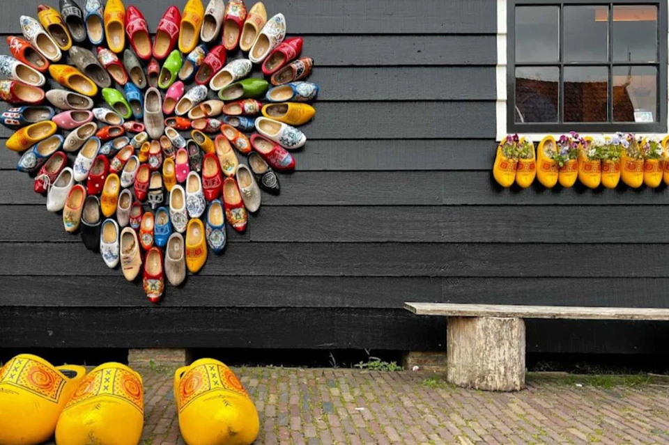 Wooden clogs arranged in a heart pattern on a black wooden wall, with a window and bench below. Large yellow clogs are on the ground.