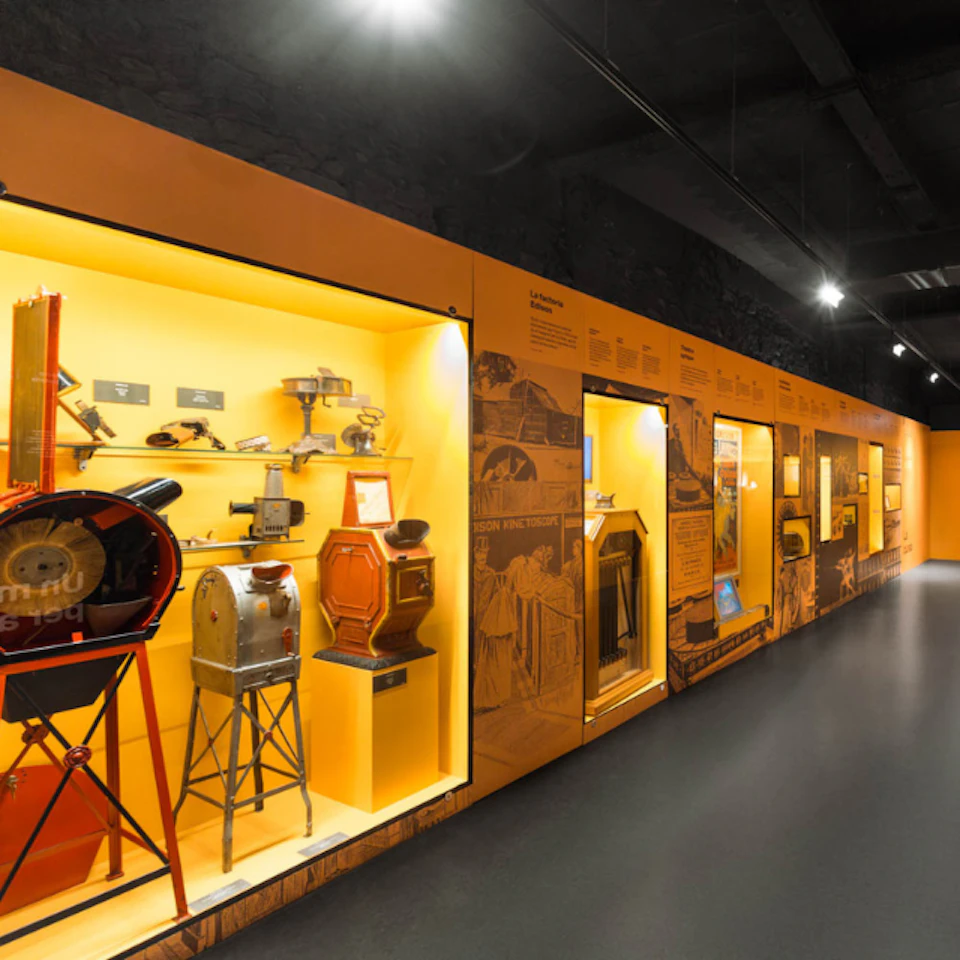A museum exhibit with vintage film projectors and cameras displayed in illuminated cases against an orange background.