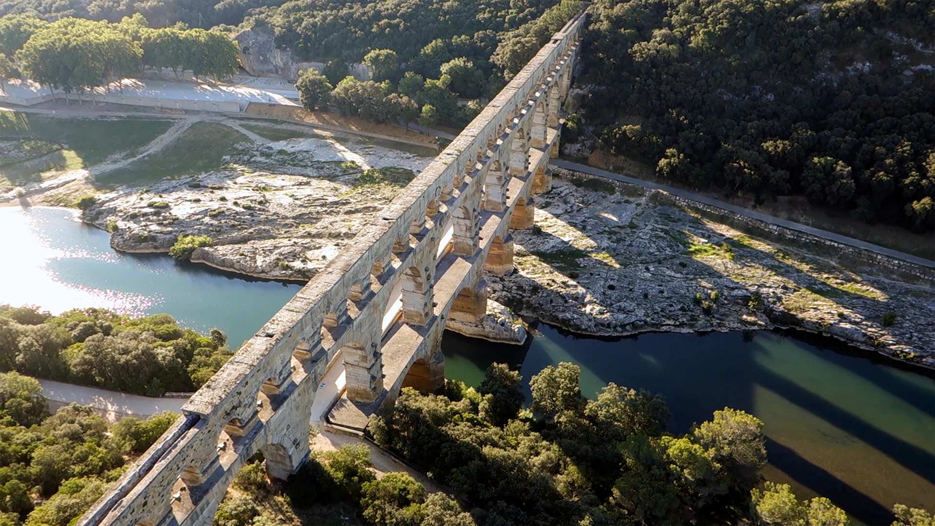 Ancient stone aqueduct spanning a river, surrounded by trees and rocky terrain.