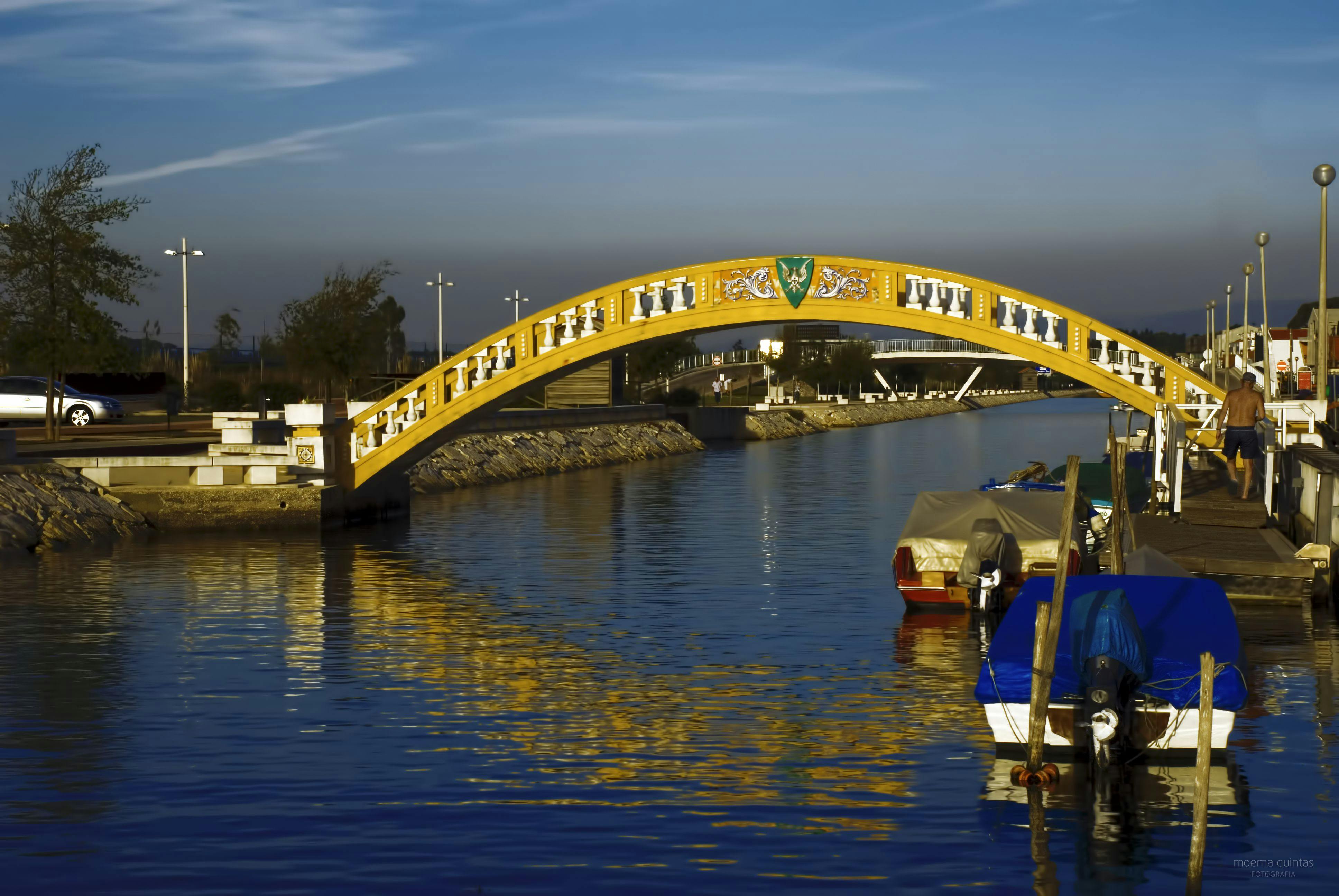 Yellow arched bridge over a calm waterway, with boats moored close by on the right. Trees and streetlights line the path beyond.