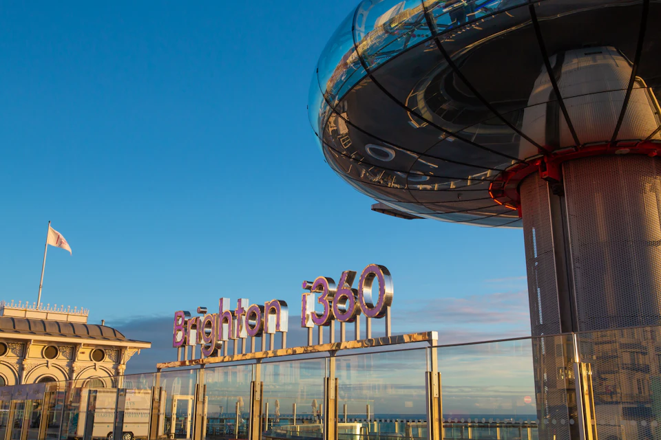 Sign reading "Brighton i360" with the observation tower extending above, set against a blue sky and ocean backdrop.