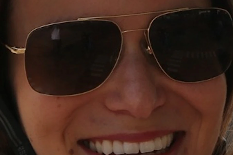 A woman wearing a red helmet and sunglasses smiles outdoors, with a modern architectural structure in the background.