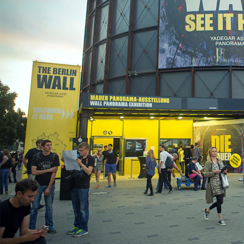 Människor samlas utanför Berlinmurens panoramautställning, med stora gula skyltar och affischer som främjar evenemanget.