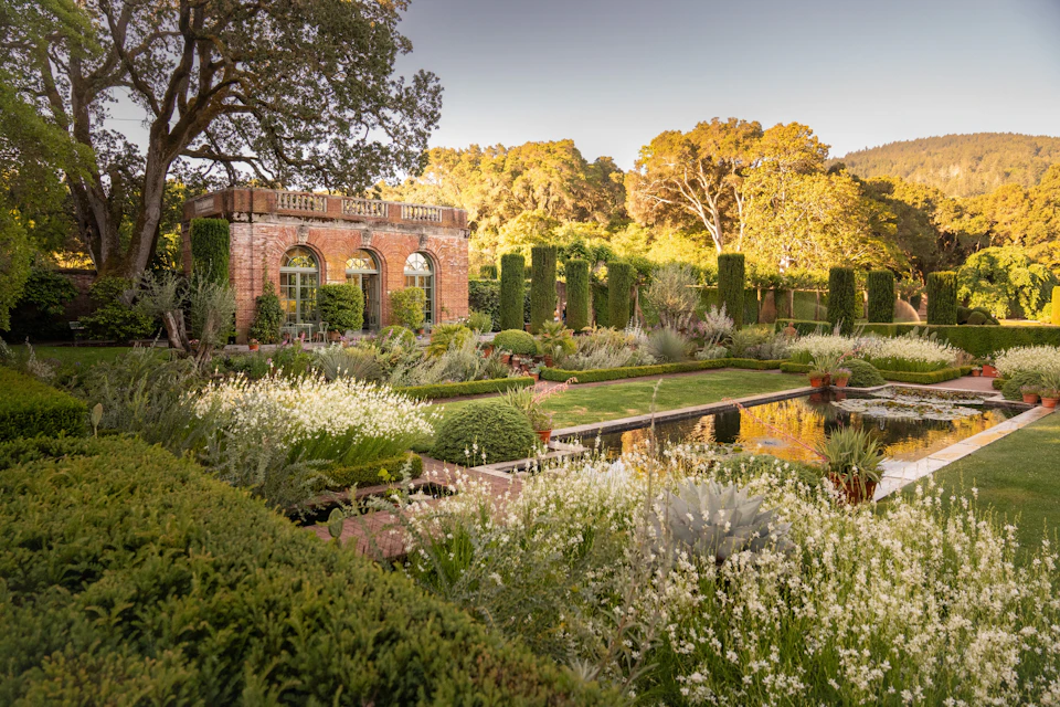 A formal garden with a brick building, trimmed hedges, flowering plants, and a reflective pond under a clear sky.